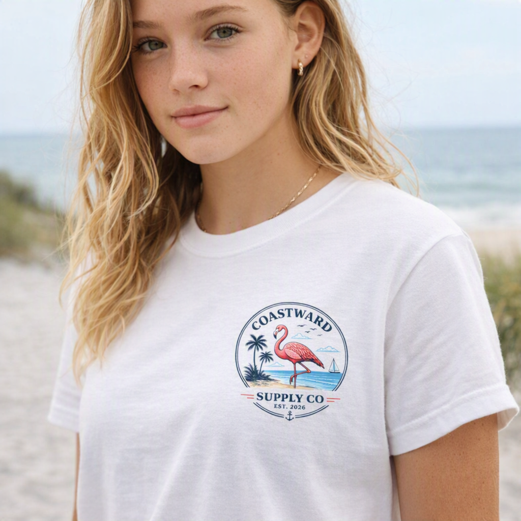 A woman wearing a white short-sleeve t-shirt with a circular 'Coastward Supply Co' flamingo graphic on the chest, set against a blurred beach background.