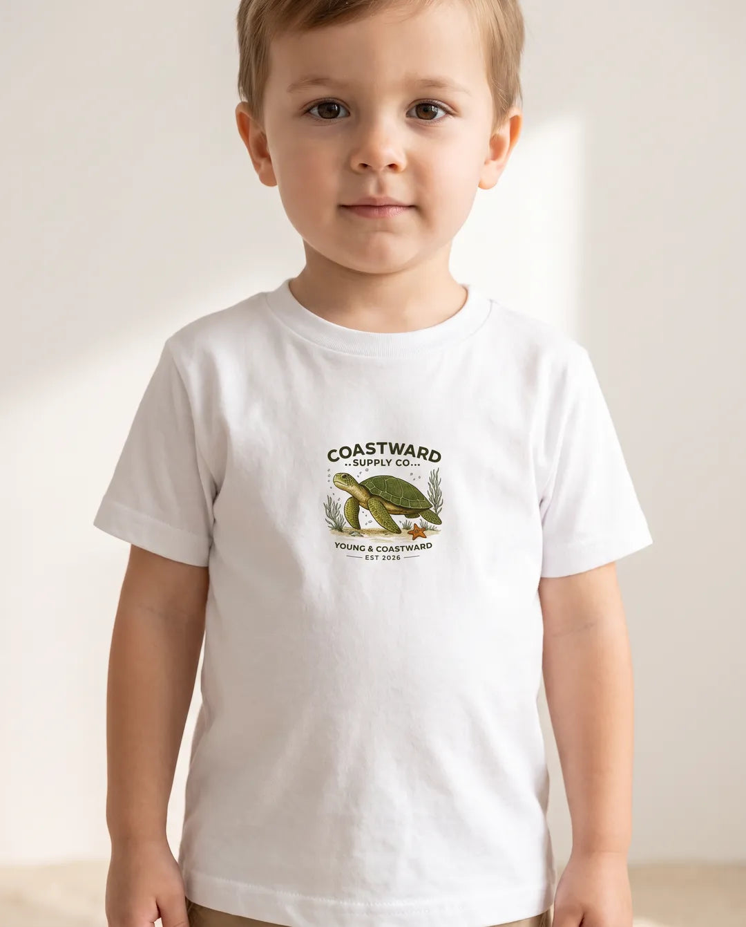 A young child wearing a white short-sleeve t-shirt with a vintage-style green turtle graphic and the text 'COASTWARD' on the chest, set against a warm, softly lit beige background.