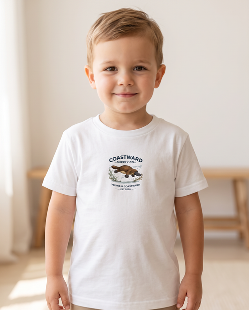 A young boy wearing a white short-sleeve t-shirt with a 'COASTWARD' graphic on the chest, standing in a brightly lit indoor room.
