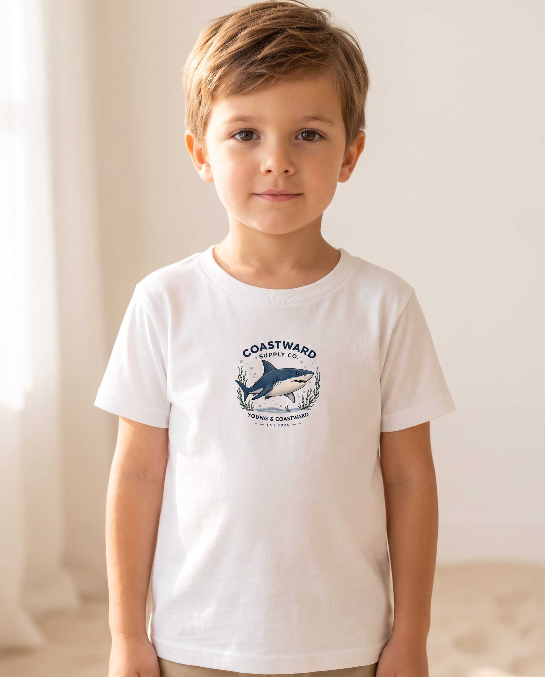 A young boy with light brown hair wearing a white short-sleeve t-shirt with a blue and gray 'Coastward' shark graphic on the chest, standing in a brightly lit indoor setting.