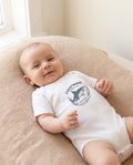 A baby lying on a beige cushion wearing a white short-sleeve bodysuit 