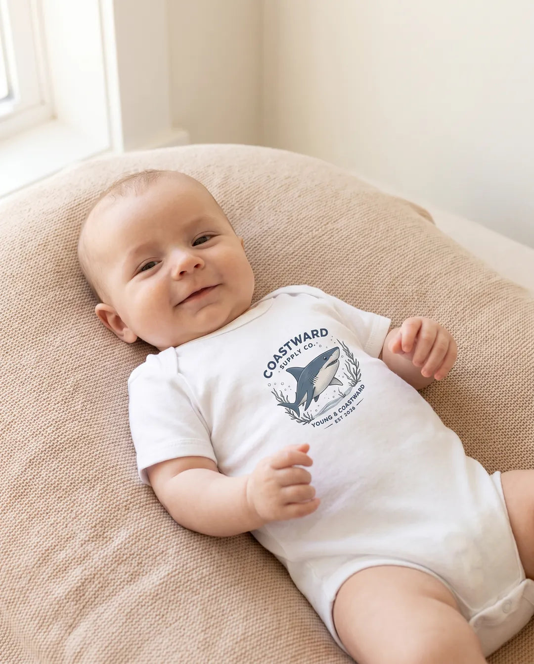 A baby lying on a beige cushion wearing a white short-sleeve bodysuit 