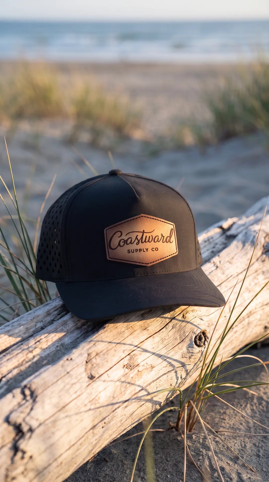 Coastward Supply Co black trucker cap with leather patch and water-drop perforated back panels, resting on driftwood at golden hour on a coastal dune